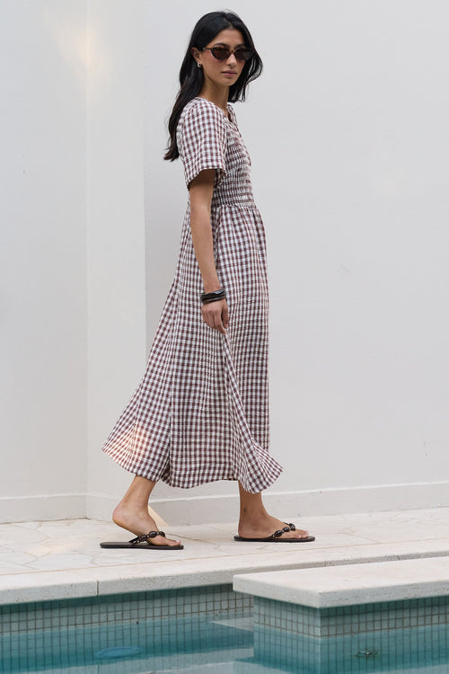 A woman wearing a brown and white gingham maxi dress and black flip-flops walks along a white tiled poolside deck next to a white wall.