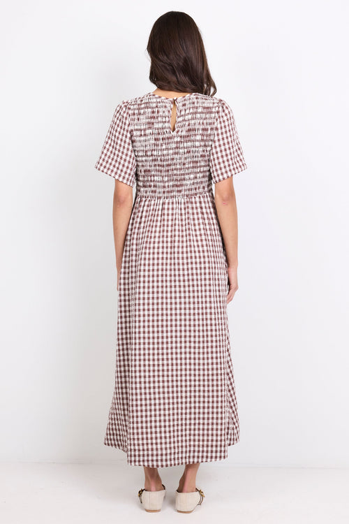 A woman viewed from the back wearing a short-sleeve midi dress with a brown and white gingham pattern and a tie detail at the back neckline, standing against a plain white studio background.