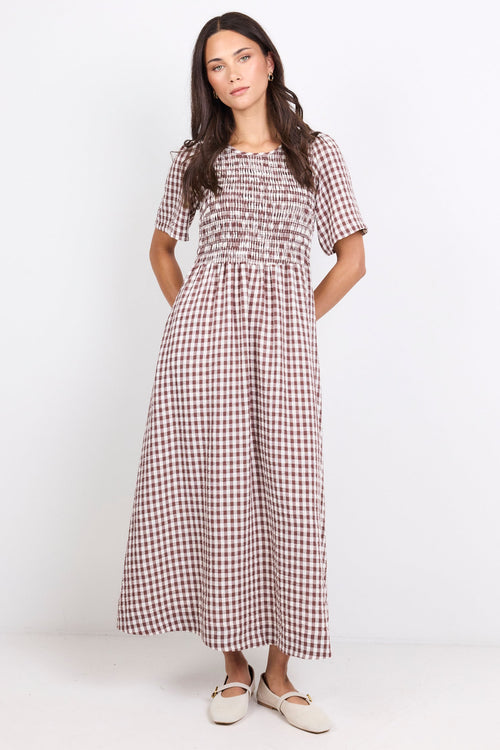 A woman modeling a short-sleeve smocked maxi dress with a brown and white gingham pattern, standing against a plain white studio background.