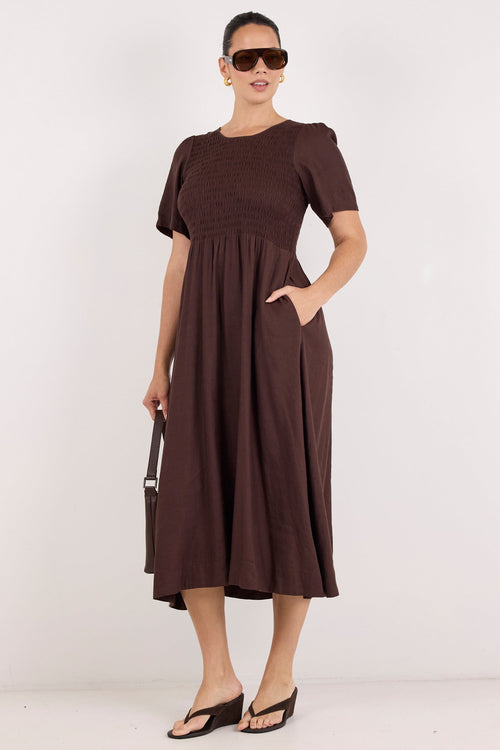 A woman modeling a dark brown short-sleeve midi dress with a gathered waist and side pockets, styled with sunglasses, a matching handbag, and black sandals against a plain white studio background.