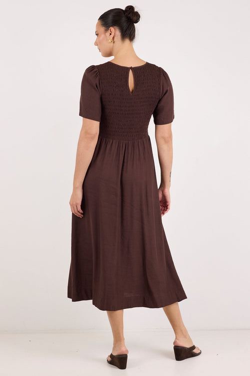 A woman modeling a dark brown midi dress from the back, featuring short puff sleeves, a smocked bodice, and a keyhole cutout at the back, set against a plain white studio background.
