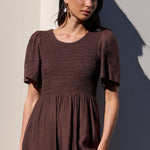 A woman wearing a dark brown smocked midi dress with short flutter sleeves, standing against a sunlit beige wall with a strong shadow.