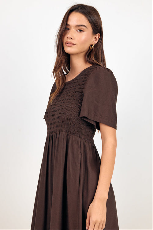 A woman modeling a dark brown smocked dress with flutter sleeves against a plain white studio background.