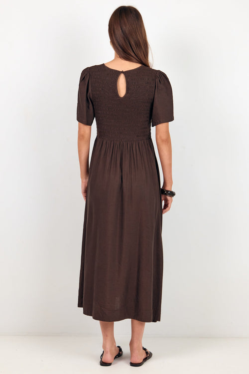 A woman viewed from the back wearing a dark brown midi dress with short puff sleeves, a smocked bodice, and a small keyhole cutout at the back neck, standing against a plain white studio background.
