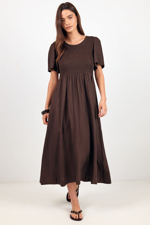A woman modeling a dark brown midi dress with short puff sleeves, a smocked bodice, and a gathered skirt, set against a plain white studio background.