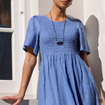 A woman wearing a smocked blue linen maxi dress with short puff sleeves, standing against a white stucco wall with a window on the left.