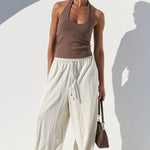 A woman models a brown halter top and cream-colored wide-leg pants against a white background with a strong shadow.