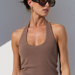 A woman stands against a white wall in a minimalist setting, wearing a brown halter top, loose light beige pants, sunglasses, and sandals, and holding a brown handbag.