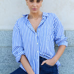 Woman wearing a blue and white striped shirt and dark jeans against a light-colored wall.