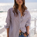 A woman wearing a brown and white vertically striped button-down shirt and blue denim jeans, standing on a sunny boardwalk with the ocean in the background.