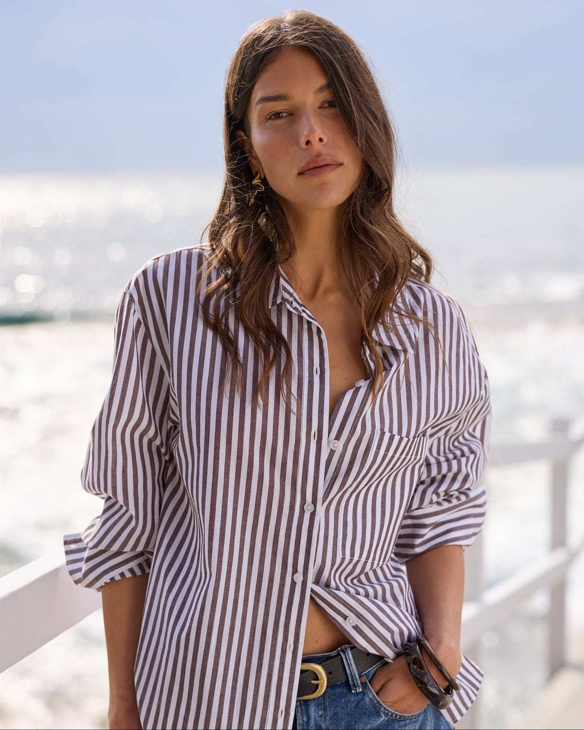 A woman wearing a brown and white vertically striped button-down shirt and blue denim jeans, standing on a sunny boardwalk with the ocean in the background.