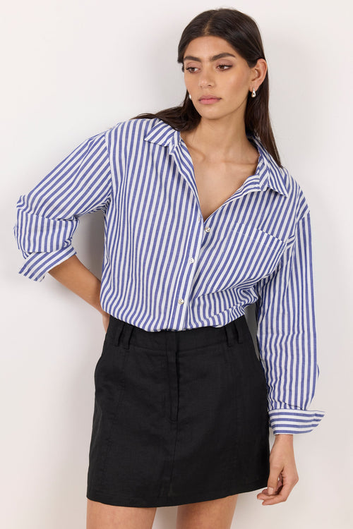 A woman modeling a blue and white vertically striped button-down shirt tucked into a black mini skirt, standing against a plain off-white background.