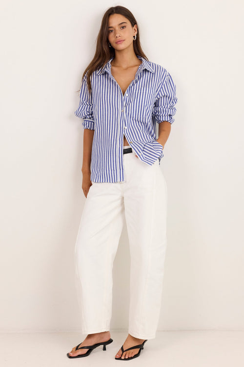A woman stands against a plain off-white wall wearing a blue and white vertically striped button-down shirt with rolled sleeves, white wide-leg cropped pants, and black strappy heeled sandals.
