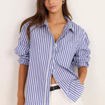 A woman modeling a blue and white vertically striped button-down shirt with white pants, standing against a plain off-white background.