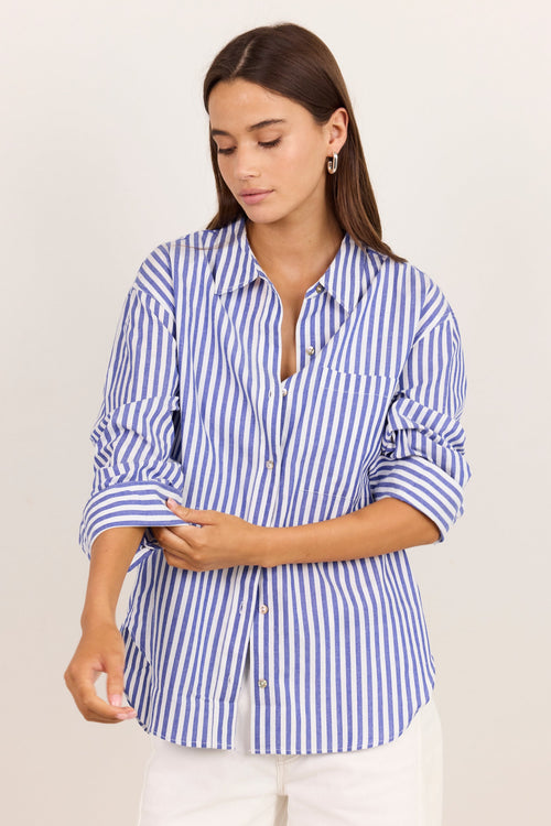 A woman wearing a blue and white vertically striped button-down shirt with rolled-up sleeves, paired with white pants, standing against a plain light beige background.