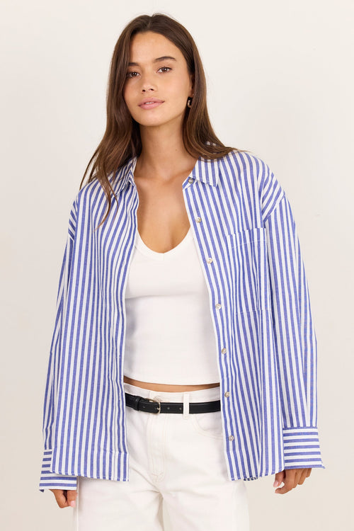 A woman modeling an oversized blue and white striped button-down shirt over a white tank top and white pants, photographed against a plain light beige studio background.