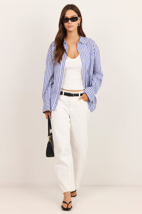 A woman modeling a blue and white striped button-down shirt over a white tank top, paired with white trousers, a black belt, black sandals, and a small black handbag, set against a plain white studio background.