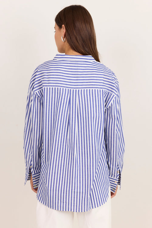 A woman viewed from the back wearing a blue and white vertically striped button-up shirt and white pants, set against a plain off-white studio background.