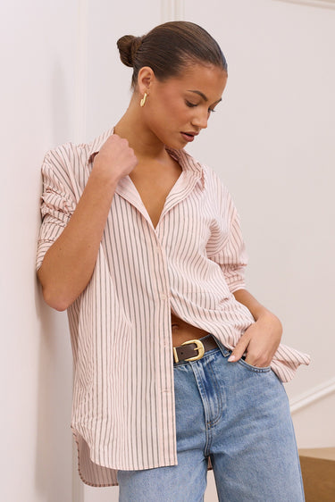 A woman wearing a light pink and white vertically striped button-down shirt and light blue jeans, leaning against a white wall on wooden stairs.
