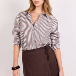 A woman modeling a brown wrap mini skirt with a tie waist and a brown-and-white vertically striped button-down shirt, standing against a plain light background.