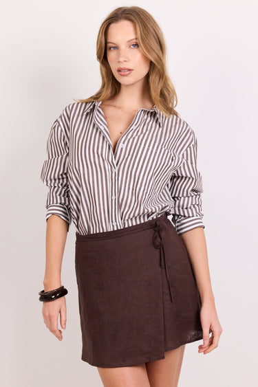A woman modeling a brown wrap mini skirt with a tie waist and a brown-and-white vertically striped button-down shirt, standing against a plain light background.