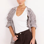 A woman modeling a casual outfit consisting of a white V-neck tank top, dark brown shorts, and a brown and white striped button-up shirt draped off her shoulders, set against a plain white background.