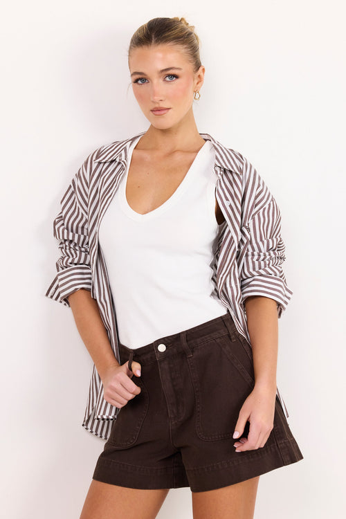 A woman modeling a casual outfit consisting of a white V-neck tank top, dark brown shorts, and a brown and white striped button-up shirt draped off her shoulders, set against a plain white background.