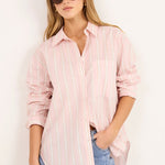 A woman modeling a light pink and white vertically striped button-down shirt, paired with light-wash blue jeans and white heeled sandals, standing against a plain white studio background.