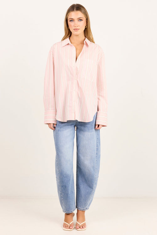 A woman stands against a plain white studio background wearing a light pink and white vertically striped button-up shirt, light-wash blue jeans, and beige strappy heels.