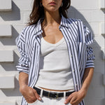 A woman wearing a navy and white vertically striped button-down shirt over a white tank top, paired with white pants and black sunglasses, posing against a white brick wall.