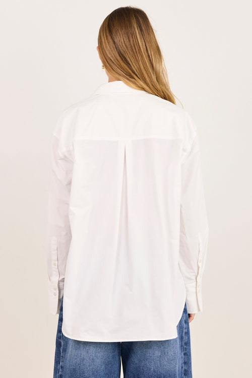 A woman viewed from behind wearing a white long-sleeve button-up shirt and blue jeans against a plain off-white studio background.