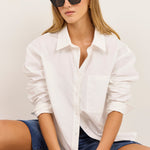 A woman sitting cross-legged wearing a white button-up shirt, blue denim shorts, black sunglasses, and black flip-flops against a plain off-white background.
