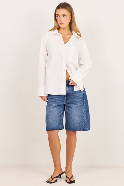 A woman modeling a white long-sleeve button-up shirt tied loosely at the waist with blue denim shorts and black sandals, standing against a plain off-white studio background.