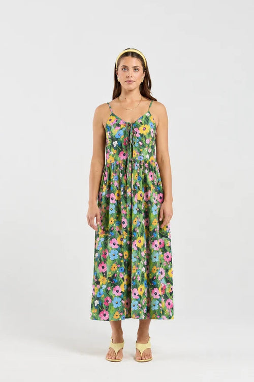 A woman modeling a sleeveless midi dress with a colorful floral print on a green background, paired with a matching headband and beige sandals, against a plain white studio background.