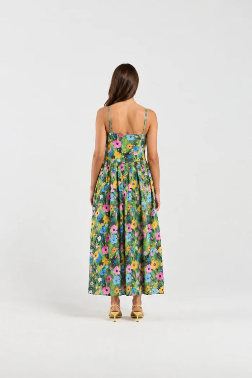 A woman viewed from behind wearing a sleeveless maxi dress with a colorful floral print on a green background, paired with tan high-heeled sandals, against a plain light gray studio backdrop.