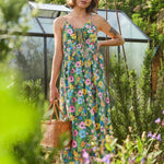 A woman stands in a sunlit garden wearing a sleeveless, tie-front maxi dress with a colorful floral print. She is holding a small woven basket bag, with a greenhouse and greenery in the background.