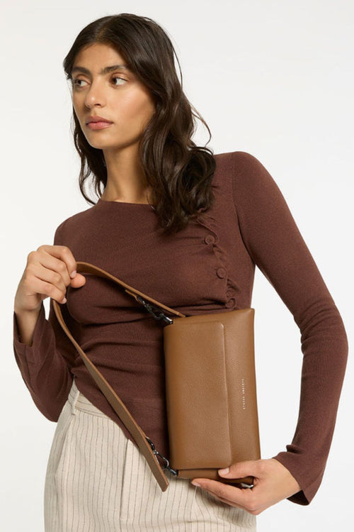 A structured camel-brown leather shoulder bag with a rectangular flap-over design and fine pebble texture. It features a detachable wide strap with black metal hardware and 