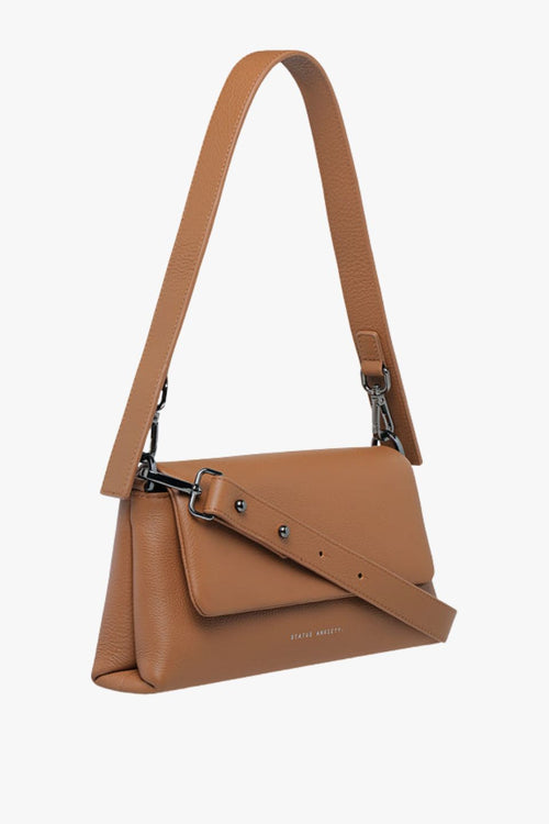 A structured camel-brown leather shoulder bag with a rectangular flap-over design and fine pebble texture. It features a detachable wide strap with black metal hardware and 