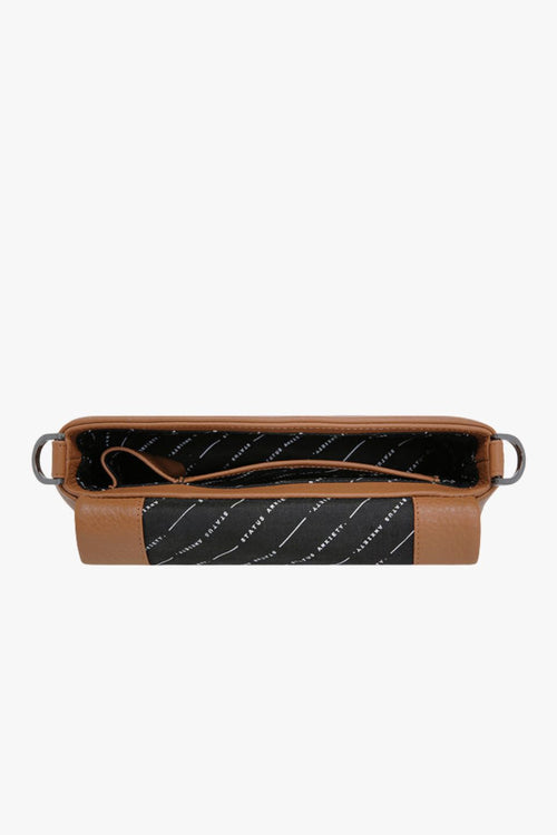 A structured camel-brown leather shoulder bag with a rectangular flap-over design and fine pebble texture. It features a detachable wide strap with black metal hardware and 