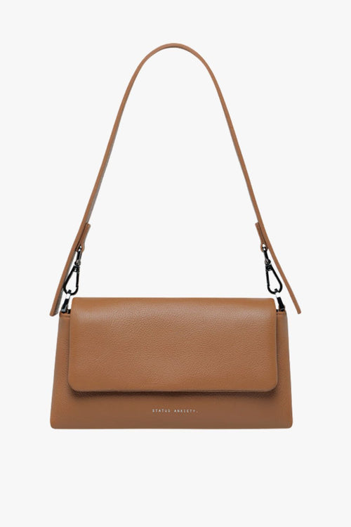 A structured camel-brown leather shoulder bag with a rectangular flap-over design and fine pebble texture. It features a detachable wide strap with black metal hardware and 