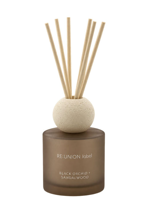 A reed diffuser bottle with a frosted brown glass base, a textured beige stone sphere, and six natural sandalwood reeds, labeled 'RE:UNION label BLACK OLEANDER + SANDALWOOD'.