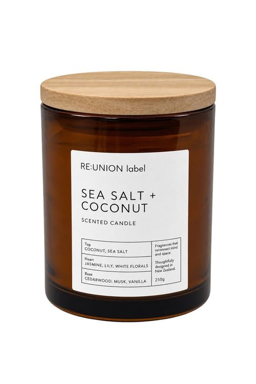 A 210g scented candle in an amber glass jar with a light wood lid, featuring a white label that reads 'RE-UNION label SEA SALT + COCONUT'.