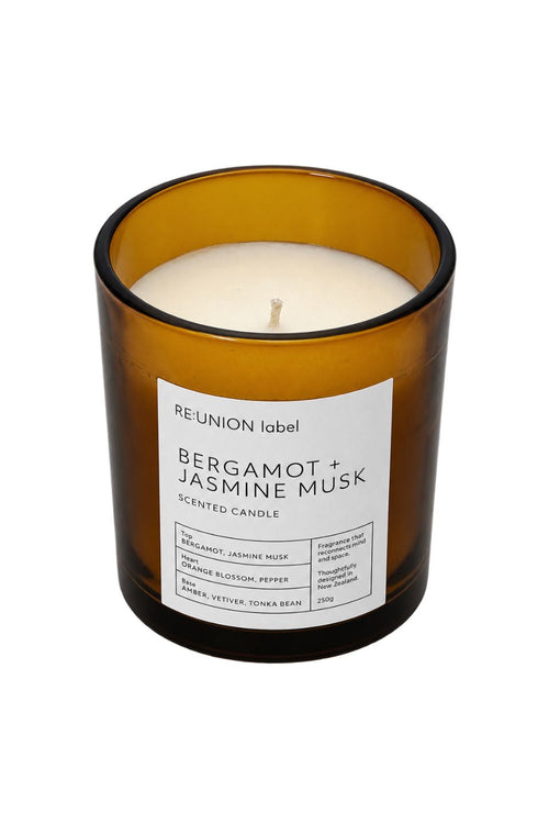 A lit scented candle in an amber glass jar with a white label reading 'REUNION label BERGAMOT + JASMINE MUSK SCENTED CANDLE'.