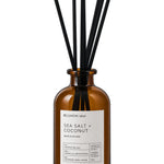 Amber glass reed diffuser bottle with a white label reading 'RE:UNION label SEA SALT + COCONUT' and eight black reed sticks, shown on a white background.