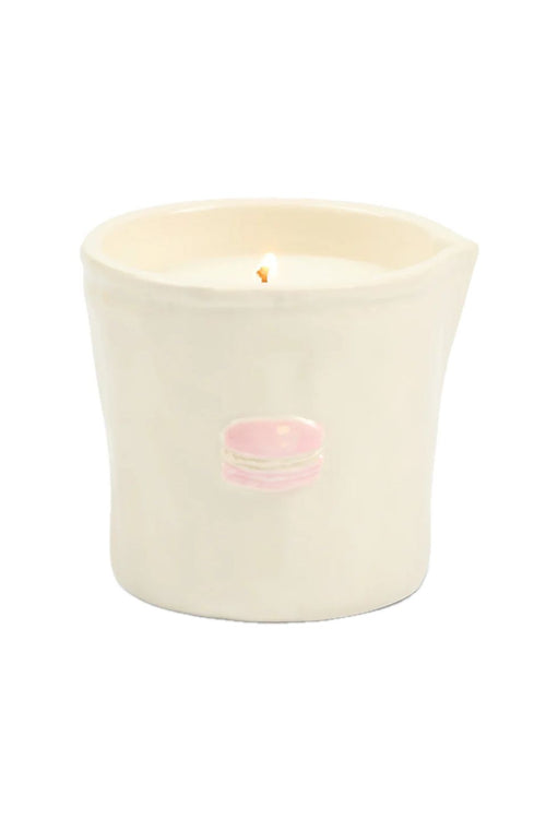 An off-white ceramic candle holder with a small pink macaron decoration on the front, containing white wax and a lit wick, set against a plain white background.
