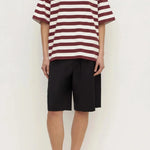 A model wearing an oversized short-sleeve t-shirt with horizontal burgundy and off-white stripes, paired with black drawstring shorts and brown sandals, standing against a plain white studio background.