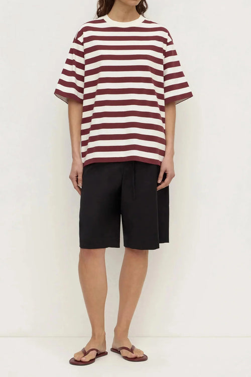 A model wearing an oversized short-sleeve t-shirt with horizontal burgundy and off-white stripes, paired with black drawstring shorts and brown sandals, standing against a plain white studio background.
