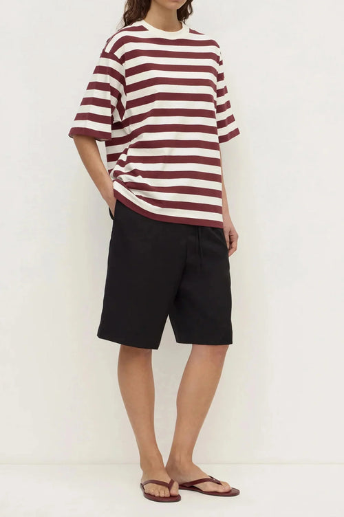 A model wearing a loose-fitting short-sleeve t-shirt with horizontal burgundy and off-white stripes, paired with black shorts and brown flip-flop sandals, standing against a plain white wall.