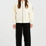 A woman stands against a plain light gray studio background wearing a cream zip-front jacket with black wide-leg pants and gray sneakers.