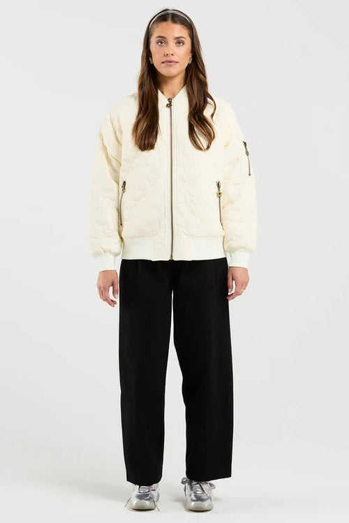 A woman stands against a plain light gray studio background wearing a cream zip-front jacket with black wide-leg pants and gray sneakers.